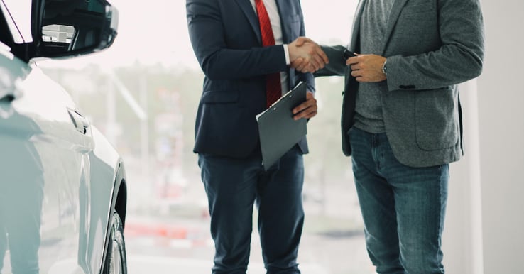 Two business men shaking hands next to a new car. One of the men holding a clipboard.